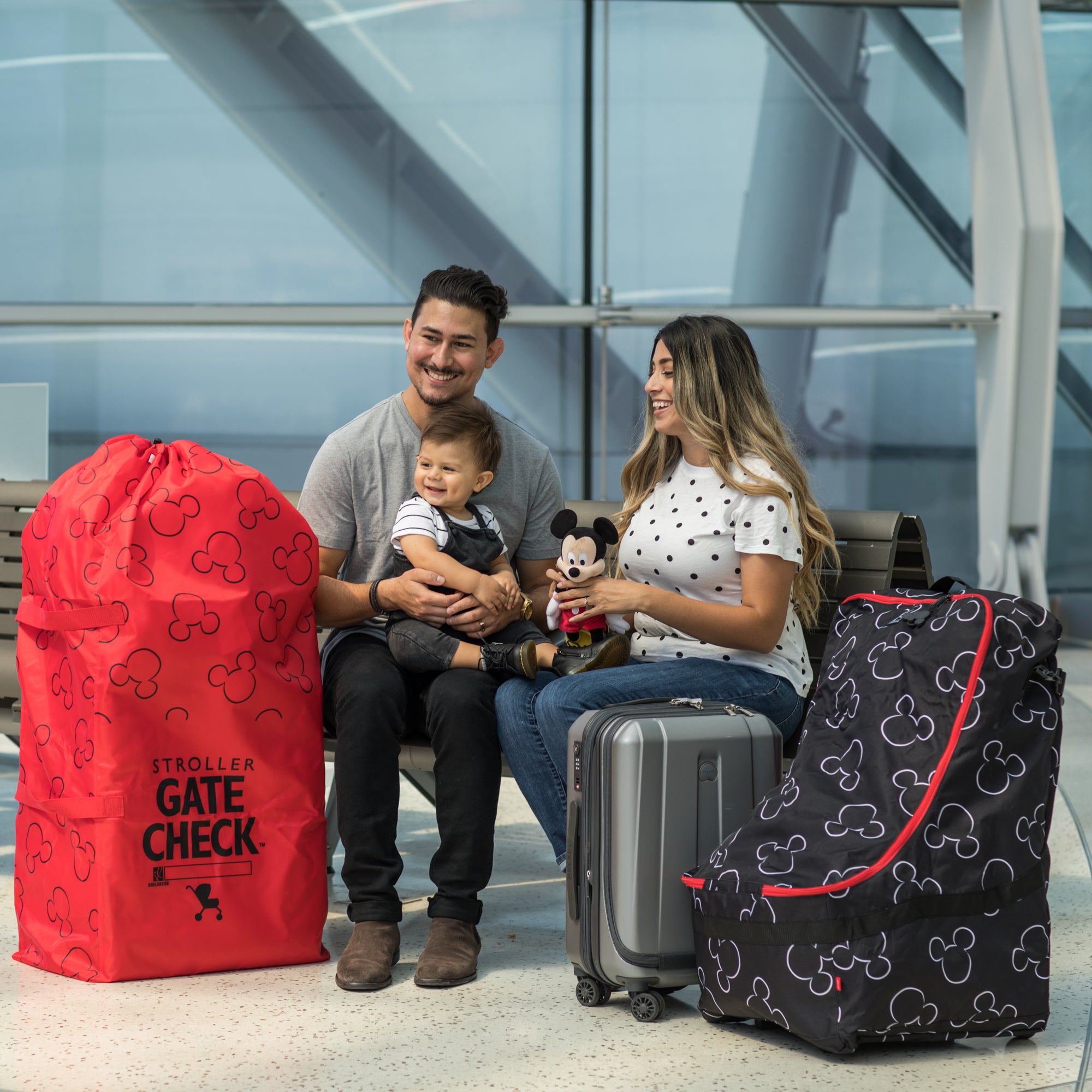 Family sitting down next to the Disney Baby Gate Check Travel Bag for Standard and Double Strollers and Disney Baby Ultimate Padded Backpack